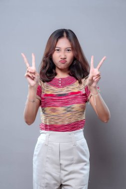 Neşeli Asyalı bir kadın, iki elini de barış işareti olarak kaldırırken komik somurtuyor. Renkli bir bluz ve açık renk pantolon giyiyor, mizah, enerji ve gençlik cazibesi sergiliyor..