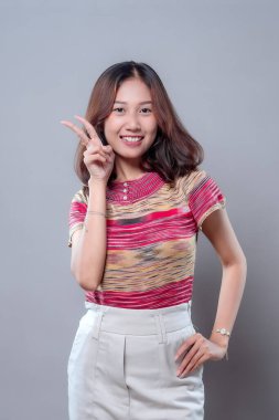 Mutlu bir Asyalı kadın güven içinde gülümserken parmaklarıyla barış işareti yapıyor, renkli çizgili bir gömlek ve beyaz pantolon giyiyor, neşeli, dostane ve pozitif enerji gösteriyor..