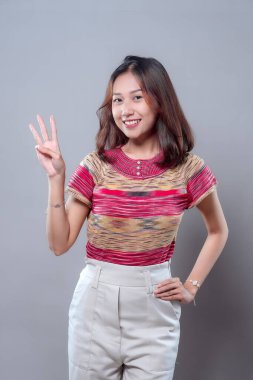 Neşeli bir Asyalı kadın üç parmak hareketi yaparken kendine güvenerek gülümsüyor. Renkli çizgili bir üst ve beyaz pantolon giyiyor, pozitifliği, kendine güveni ve gençlik enerjisini ifade ediyor..