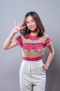 Mutlu bir Asyalı kadın dört parmak hareketi yaparken ışıl ışıl gülümsüyor. Renkli çizgili bir gömlek ve beyaz pantolon giyiyor. Pozitif, kendine güvenen ve gençlik enerjisini dostça bir duruşla ifade ediyor..