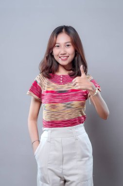 Kendine güvenen Asyalı bir kadın başparmak hareketi yaparken ışıl ışıl gülümsüyor. Renkli desenli bir gömlek ve beyaz pantolon giyiyor, pozitifliğini, başarısını, onayını ve modern özgüvenini ifade ediyor..