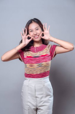 Neşeli, genç Asyalı bir kadın iki kere tamam el hareketi yaparken ışıl ışıl gülümsüyor. Renkli çizgili bir bluz ve beyaz pantolon giyiyor, pozitifliği, dostluğu ve modern güveni ifade ediyor..