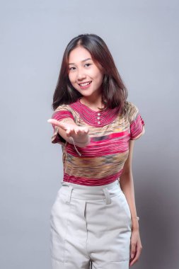 Dost canlısı genç bir Asyalı kadın elini ileri uzatırken sıcak bir şekilde gülümsüyor. Renkli çizgili bir bluz ve beyaz pantolon giyiyor. Nezaket, davetiye ve olumlu bir bağlantı gösteriyor..
