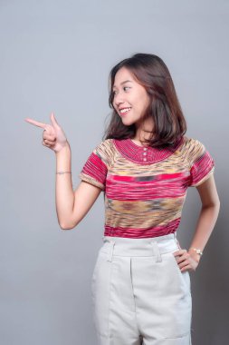 Neşeli, genç Asyalı bir kadın, kendine güvenle gülümserken, renkli bir bluz ve açık renk pantolon giyer, yönü, fikir sunumunu ve pozitif iletişimi sembolize eder..