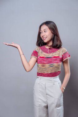 Genç Asyalı bir kadın, elleri açık bir şekilde gülümserken, renkli bir bluz ve beyaz pantolon giyiyor, pozitifliği, güveni ve dostça iletişimi temsil ediyor..