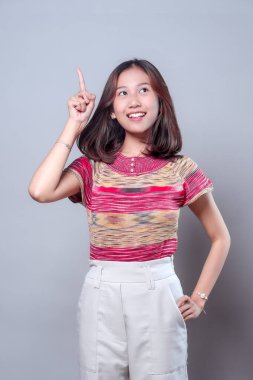 Genç Asyalı bir kadın işaret parmağıyla yukarıya doğru gülümserken renkli bir bluz ve açık renk pantolon giyerek yaratıcılık, güven ve pozitif ilham gösteriyor..