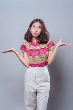 Şaşırmış bir ifadeyle genç bir Asyalı kadın omuzlarını silkiyor ve iki elini kaldırıyor. Renkli bir bluz ve beyaz pantolon giyiyor. Günlük olaylarda kafa karışıklığını, şüpheyi ya da belirsizliği temsil ediyor..