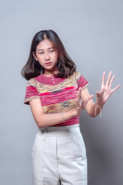 Tedbirli bir ifadeye sahip genç bir Asyalı kadın iki elini birden kaldırıyor. Renkli çizgili gömlek ve beyaz pantolon giyiyor. Korkuyu, kaçınmayı veya iletişimde tereddüt etmeyi simgeliyor..