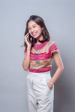 Akıllı telefonuyla konuşurken gülümseyen neşeli bir Asyalı kadın, renkli bir bluz ve beyaz pantolon giyiyor. Onun parlak ifadesi arkadaşlığı, güveni ve olumlu iletişimi yansıtıyor..