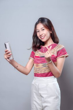 Zeki telefonuyla selfie çekerken gülümseyen neşeli bir Asyalı kadın küçük bir kalp hareketi gösteriyor, sevgi, neşe ve modern yaşam tarzında pozitif bir iletişim gösteriyor..