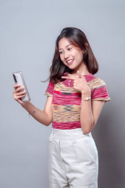 Mutlu bir Asyalı kadın selfie çekerken gülümsüyor ve akıllı telefonuna işaret ediyor, kendine güvenini ve heyecanını ifade ediyor, modern yaşam tarzını, iletişimini ve sosyal bağlantısını temsil ediyor..