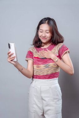 Asyalı bir kadın gözleri kapalı gülümsüyor, bir elini akıllı bir telefon tutarken göğsüne koyuyor, minnettarlığını ve barışçıl bir duyguyu sakin ve modern bir görünümle ifade ediyor..