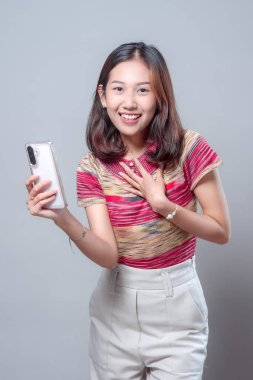 Akıllı bir telefonla gülümseyen ve tek eliyle göğsüne dokunan Asyalı bir kadın mutluluk ve minnettarlık gösteriyor. Renkli çizgili bir bluz ve açık renk pantolon giyiyor..