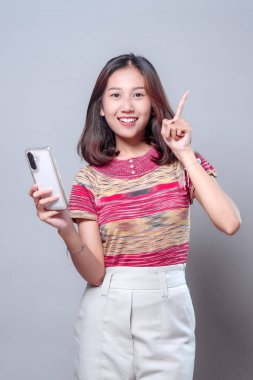 Akıllı bir telefonla gülümseyen Asyalı bir kadın yukarı doğru işaret ederek parlak bir fikir gösteriyor. Renkli çizgili bir bluz ve açık renk pantolon giyiyor, pozitifliğini ve kendine güvenini gösteriyor..