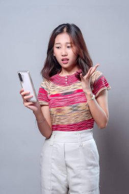 Şok olmuş ve kafası karışmış bir ifadeyle akıllı telefonuna bakan Asyalı bir kadın. Renkli çizgili bir bluz ve açık renk pantolon giyiyor. Modern bir yaşam tarzına olan inancını ya da hayal kırıklığını ifade ediyor..