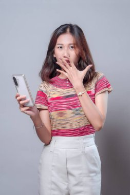 Endonezyalı bir Asyalı kadın akıllı telefon tutarken ağzını eliyle kapatırken, renkli örgü bir bluz ve temiz, nötr bir düzende beyaz pantolon giyerken şaşırmış görünüyor..