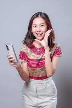 Endonezyalı bir Asyalı kadın akıllı telefonu tutarken parlak bir şekilde gülümsüyor. Elleriyle bir jest yapıyor. Renkli örgü bir bluz ve beyaz bir pantolon giyiyor..