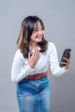 Akıllı telefonunu tutarken sıcacık gülümseyen neşeli bir Asyalı kadın video görüşmesinden hoşlanıyor gibi görünüyor. Beyaz bir bluz ve kot etek giyiyor, mutluluğu, arkadaşlığı ve bağlantıyı ifade ediyor..