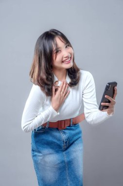 Akıllı telefonunu tutarken gülümseyen ve göğsüne minnettarlıkla dokunan neşeli, genç bir Asyalı kadın. Beyaz bir bluz ve kot etek giyiyor, mutluluğu, nezaketi ve samimiyeti ifade ediyor..