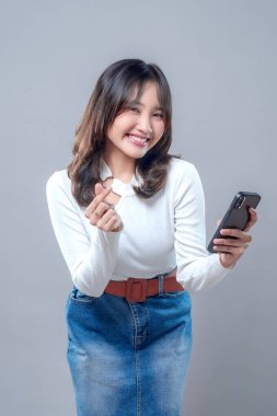 Neşeli, genç Asyalı bir kadın, parmaklarıyla kalp hareketi yaparken ve akıllı telefonunu tutarken ışıl ışıl gülümsüyor. Uzun kollu beyaz bir bluz ve kot etek giyiyor. Sevgisini, neşesini ve pozitifliğini ifade ediyor..