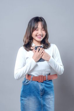 Genç bir Asyalı kadın göğsüne yakın bir akıllı telefonu tutarken gözleri kapalı nazikçe gülümsüyor. Uzun kollu beyaz bir bluz ve kot etek giyiyor. Minnettarlığını, sevgisini ve mutluluğunu ifade ediyor..