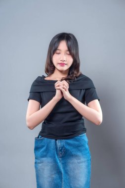 Asyalı bir kadın, gözleri kapalı ve sakin bir ifadeyle ellerini bir arada tutuyor, siyah bir bluz ve kot etek giyiyor, huzuru, farkındalığı ve zarif güzelliği huzurlu bir anda gösteriyor..