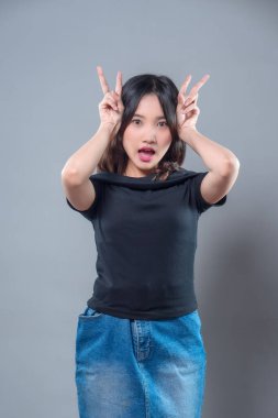 Uzun saçlı neşeli bir Asyalı kadın parmakları tavşan kulağı şeklinde neşeli bir jest yapıyor. Siyah bir gömlek ve kot etek giyiyor, sürpriz, eğlence ve gençlik enerjisini ifade ediyor..