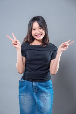İki eliyle iki barış işareti gösterirken parlak bir şekilde gülümseyen genç bir Asyalı kadın. Siyah bir gömlek ve kot etek giyiyor, kendine güveni, neşesi, arkadaşlığı ve gençlik enerjisini ifade ediyor..