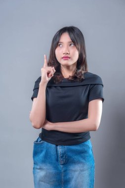 Genç Asyalı bir kadın tek parmakla yukarıya doğru işaret ederken düşünceli görünüyor, siyah gömlek ve kot etek giymiş, odaklanmış bir ifadeyle bir an veya yeni bir fikir ifade ediyor..