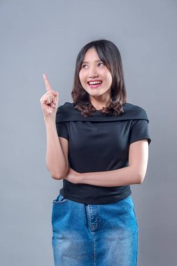 Çekici bir Asyalı kadın tek parmakla yukarıya doğru gülümserken neşeli bir ifade ve modern kıyafetlerle pozitif bir fikir konsepti sergiliyor..
