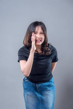 Asyalı bir kadın acı dolu bir ifadeyle yanağına dokunuyor, diş ağrısından rahatsızlık duyuyor. Görüntü diş ağrısını, oral sağlık sorunlarını ve tıbbi tedavi veya bakım ihtiyacını temsil ediyor..