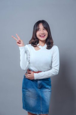 Neşeli, genç Asyalı bir kadın yukarı bakıp barış işareti gösterirken ışıl ışıl gülümsüyor. Uzun kollu beyaz bir bluz ve kot etek giyiyor, pozitifliğini ve özgüvenini ifade ediyor..