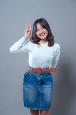 Barış işareti ile poz verirken gülümseyen mutlu genç bir Asyalı kadın. Uzun kollu beyaz bir bluz ve kot etek giyiyor. Kendine güvenini, dostluğunu ve pozitif enerjisini gösteriyor..
