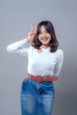 Yüzünde barış işareti ile poz veren neşeli, genç bir Asyalı kadın. Uzun kollu beyaz bir bluz ve kot etek giyiyor. Cazibesini, özgüvenini ve pozitif enerjisini gösteriyor..