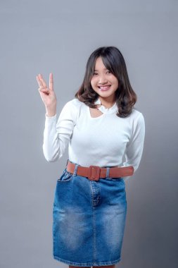 Üç parmaklı bir jest yaparken kendine güvenen neşeli, genç bir Asyalı kadın. Beyaz bir bluz ve kahverengi kemerli kot etek giyiyor. Arkadaşlığını, pozitifliğini ve modern çekiciliğini ifade ediyor..