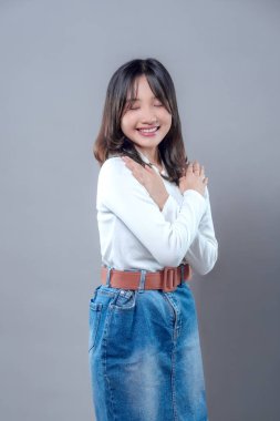 Gözleri kapalı gülümseyen, beyaz bir kazak ve kot etek giyen, huzur, rahatlık ve kendini sevme gösteren, kollarını göğsüne çarpıştıran neşeli, genç bir Asyalı kadın..