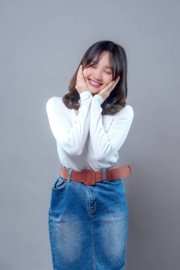 Gözleri kapalı gülümseyen, elleri yanaklarında beyaz bir bluz ve kot etek giyen sakin bir Asyalı kadın..