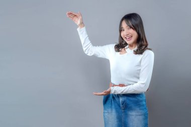 Neşeli, genç Asyalı bir kadın iki eliyle işaret ederken gülümsüyor. Yanındaki boşluğu göstermek için. Uzun kollu beyaz bir bluz ve kot etek giyiyor..
