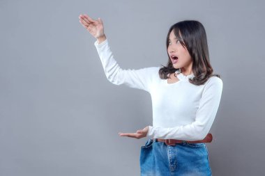 Şaşırmış bir ifadeyle genç bir Asyalı kadın, her iki eliyle, beyaz uzun kollu bluz ve kot etek giyen, boş bir alan sunmak için işaret ederken hayrete düşüyor..