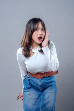 Şaşırmış bir ifadeyle Asyalı bir kadın, ağzı açık ve eli yüzüne yakın, beyaz uzun kollu bluz ve kahverengi kemerli kot etek giyiyor, duygularını ve merakını gösteriyor..