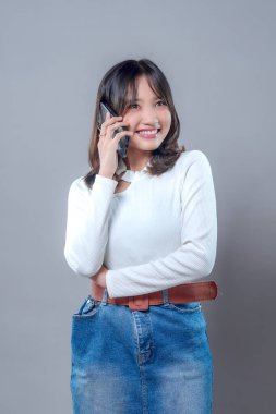 Akıllı telefonuyla konuşurken gülümseyen ve neşeli bir ifadeyle yüzünü çeviren genç bir Asyalı kadın. Uzun kollu beyaz bir bluz ve kot etek giyiyor. Kendine güveni ve pozitifliği var..