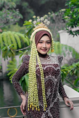 Endonezyalı bir kadın geleneksel gelin kebaya bürünmüş kahverengi tesettür ve altın tacıyla, zarif bir şekilde açık havada gülümsüyor, yasemin çelenkleri, dantel eldivenler ve detaylı boncuklarla süslenmiş..