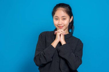 Elleri çenesinin altına sıkışmış gülümseyen genç bir kadın, kameraya bakarken samimi ve samimi görünüyor, mavi arka planda sıcak ve samimi bir ifade sergiliyor..