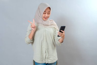 Tesettürlü genç Asyalı kadın gülümseyerek selfie çekiyor ve parmaklarıyla barış işareti gösteriyor. Neşeli ifade, günlük tarz, gri arkaplanda izole..