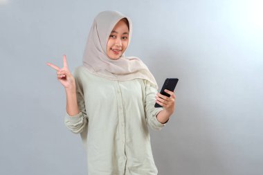 Tesettürlü genç Asyalı kadın akıllı telefonuna bakıp barış işareti gösterirken kendine güvenerek gülümsüyor. Olumlu yüz ifadesi, modern ve neşeli ruh hali, izole edilmiş..