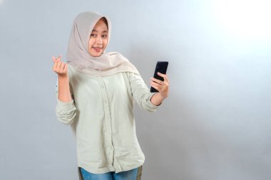 Tesettürlü neşeli, genç Asyalı kadın selfie çekerken gülümsüyor ve parmak kalp hareketi yapıyor. Parlak yüz ifadesi, kendinden emin ve arkadaş canlısı tavır, stüdyo çekimi..