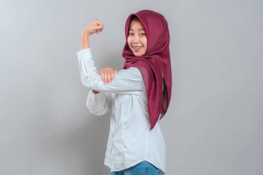 Kestane rengi tesettürlü kendine güvenen genç bir kadın kol kaslarını esnerken gülümsüyor, gücünü ve gücünü gösteriyor. Onun mutlu ve gururlu ifadesi pozitiflik ve kararlılığı ifade eder..