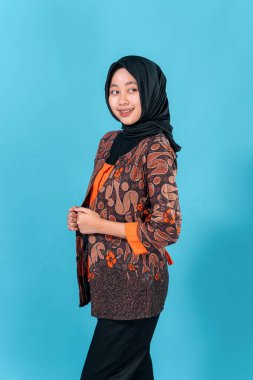Gülümseyen, tesettürlü genç Asyalı kadın neşeli ve kendinden emin bir ifade takınıyor, geleneksel batik kıyafetler giyiyor, sağlam mavi bir arka plana karşı duruyor..