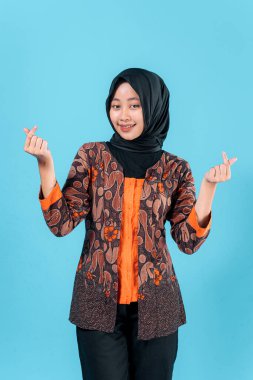 Tesettürlü ve geleneksel batik kıyafetli gülümseyen genç Asyalı kadın stüdyo resminde neşe ve dostluğu ifade eden bir parmak hareketi yapıyor..
