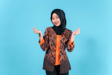 Tesettürlü ve geleneksel batik kıyafetli gülümseyen genç Asyalı kadın stüdyo resminde neşe ve dostluğu ifade eden bir parmak hareketi yapıyor..